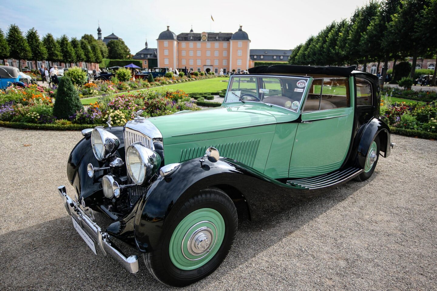 Klassisches Auto in einem Garten mit Blumen und einem historischen Gebäude im Hintergrund.