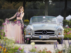 Frau in floralem Kleid posiert neben einem silbernen Mercedes Cabrio in einem Garten.