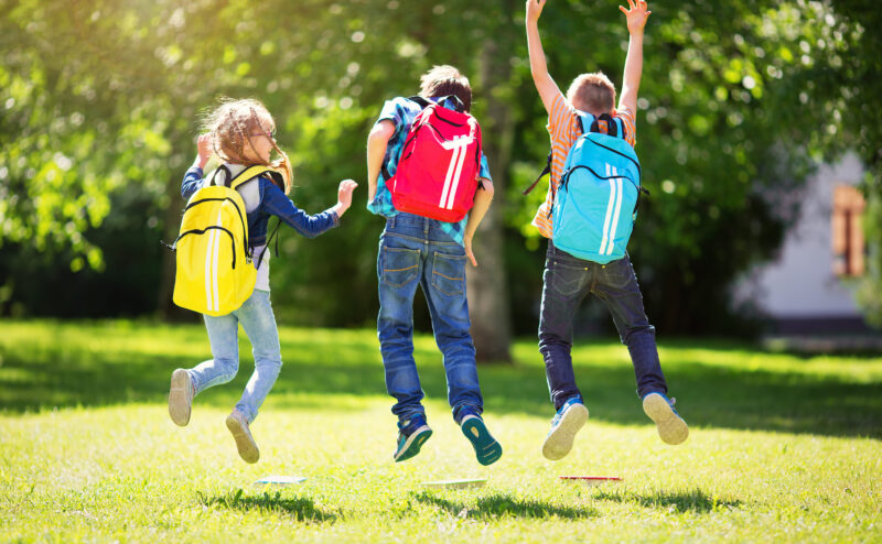 Fröhliche Schulkinder mit bunten Rucksäcken springen voller Freude auf einer sonnigen Wiese. Die Kinder tragen verschiedene farbenfrohe Kleidung, darunter Jeans und T-Shirts, und ihre Rucksäcke in Gelb, Rot und Blau heben sich lebhaft vom satten Grün des Rasens und der Bäume im Hintergrund ab. Die Szene vermittelt eine Atmosphäre von Freiheit, Freude und unbeschwerter Kindheit in einer natürlichen Umgebung.