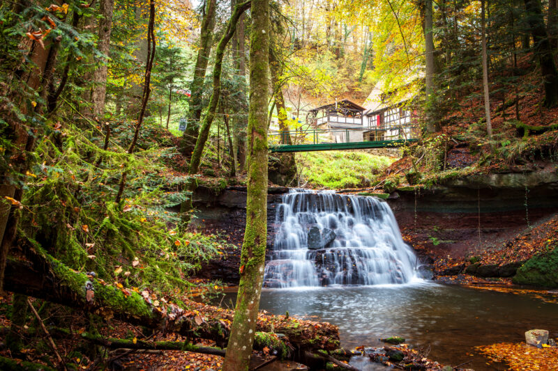 Ein Wasserfall umgeben von Bäumen und einem Holzhaus im Hintergrund.