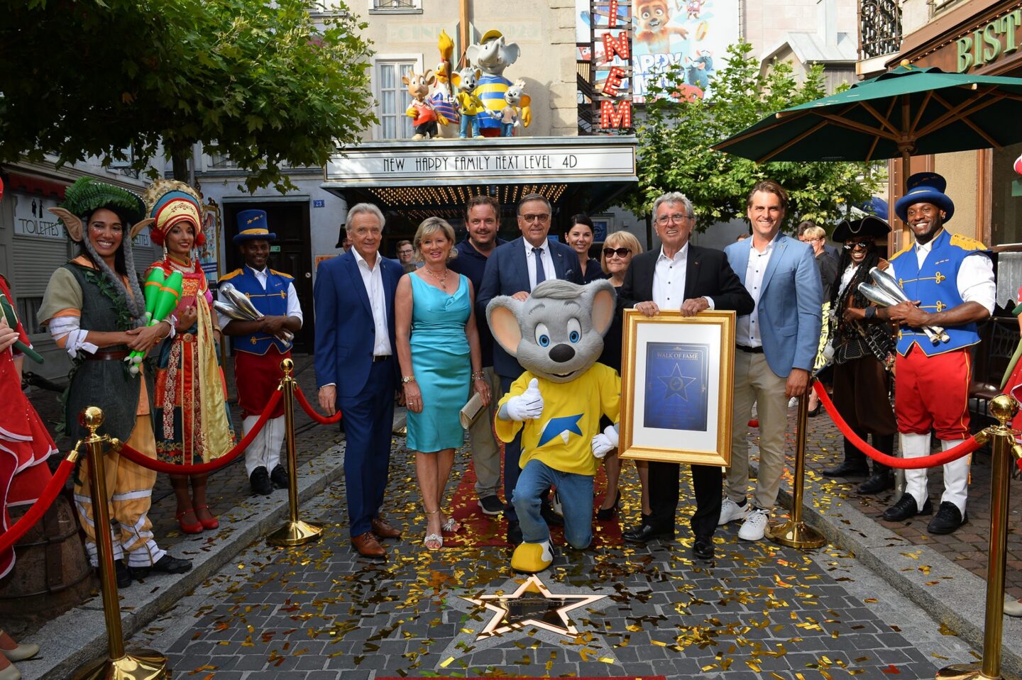 Gruppenfoto mit einem Maskottchen und mehreren Personen in festlicher Kleidung vor einem Freizeitpark.
