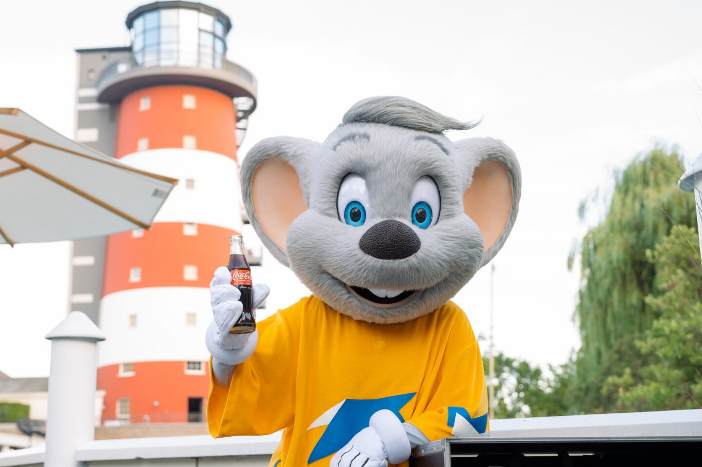 Ein Maskottchen in einem gelben Shirt hält eine Cola-Flasche vor einem Leuchtturm.