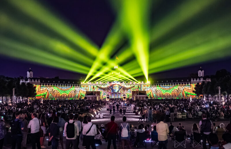 Große Menschenmenge bei einer Lichtshow mit bunten Strahlen und Projektionen.