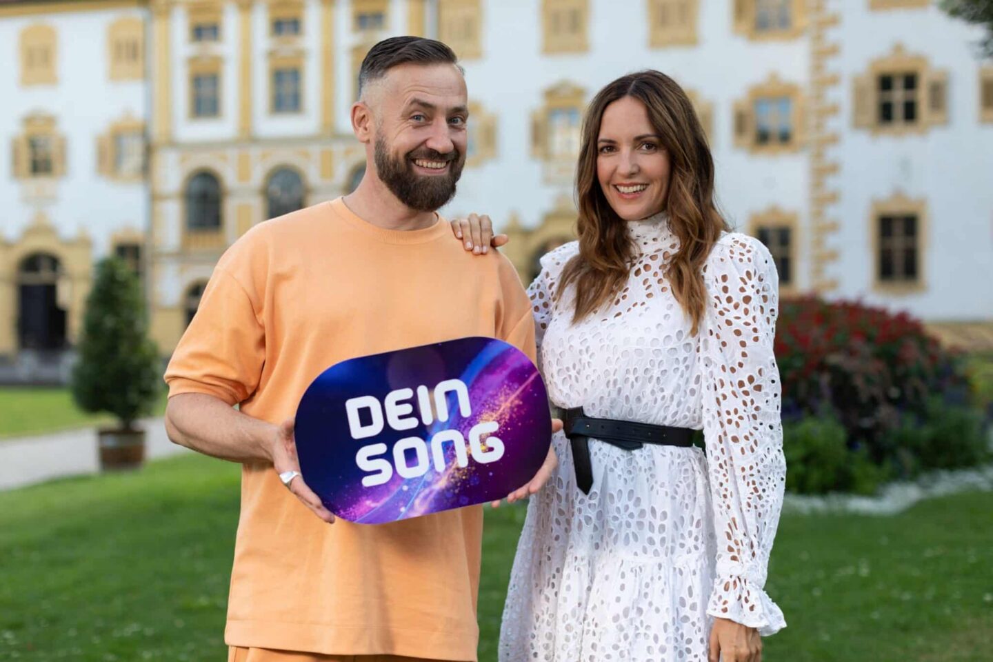 Zwei Personen halten ein Schild mit der Aufschrift 'DEIN SONG' in einem Garten.
