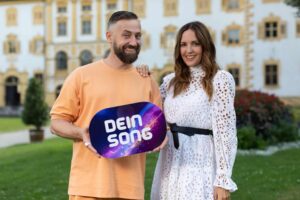 Zwei Personen halten ein Schild mit der Aufschrift 'DEIN SONG' in einem Garten.
