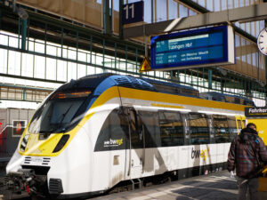 Zug am Bahnhof mit Anzeigetafel und Fahrgästen.