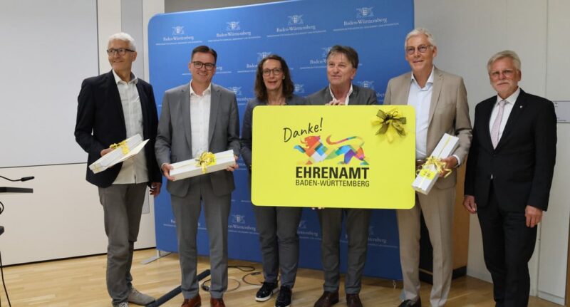 Sechs Personen stehen vor einer Wand mit dem Schriftzug 'Ehrenamt Baden-Württemberg'.