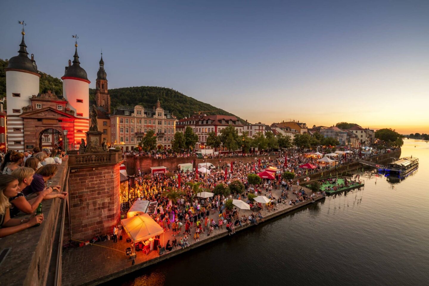 Blick auf ein Fest am Fluss mit Menschenmengen und bunten Ständen bei Sonnenuntergang.