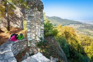 Zwei Personen liegen an einer alten Steinmauer mit Blick auf eine grüne Landschaft.