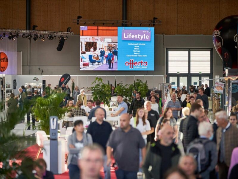 Menschenmenge auf einer Messe mit einem großen Bildschirm im Hintergrund.