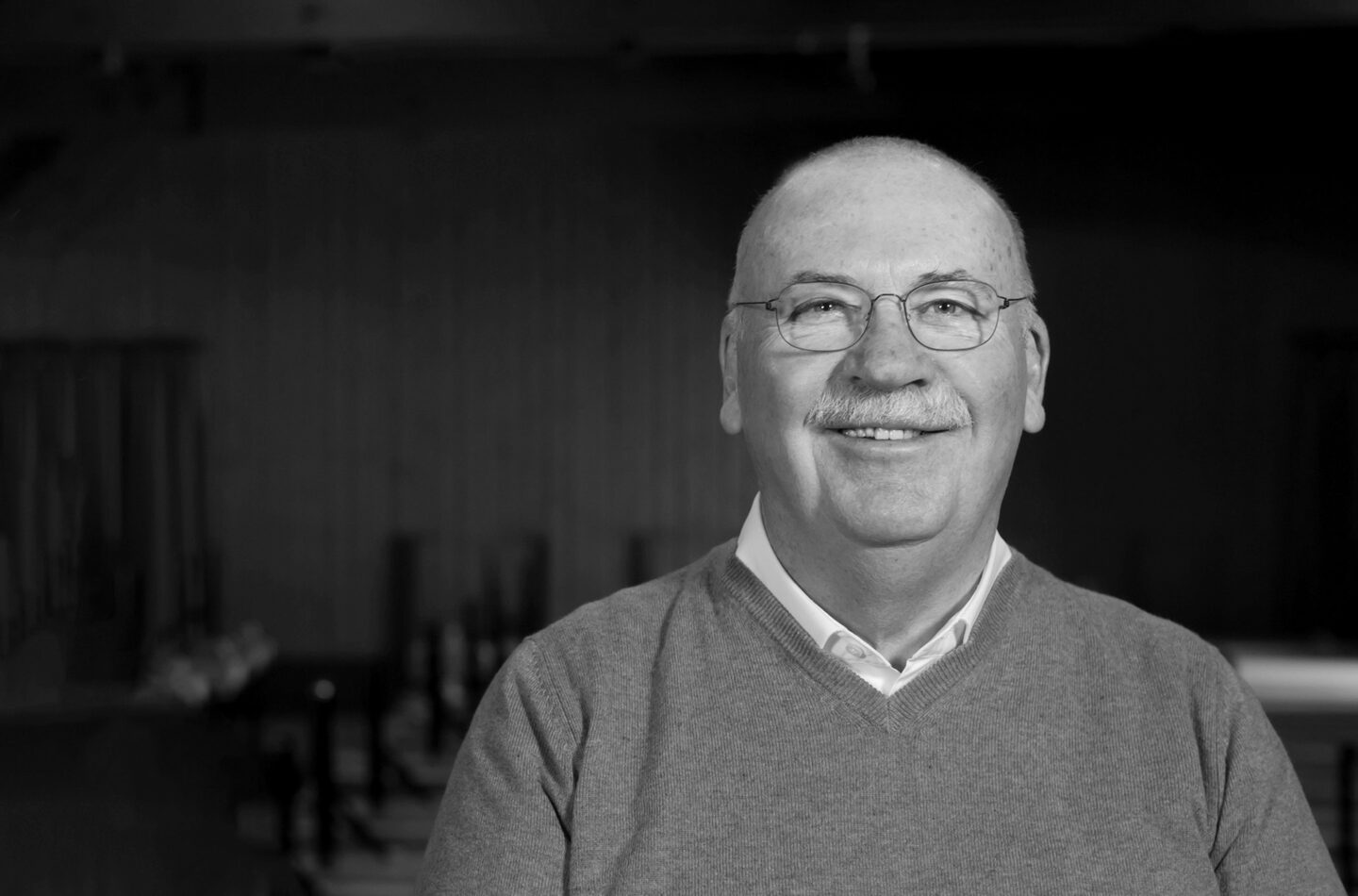 Lächelnder Mann mit Brille und grauem Pullover in einem dunklen Raum.