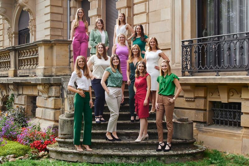 Gruppenfoto von Frauen in eleganter Kleidung auf einer Treppe vor einem historischen Gebäude.
