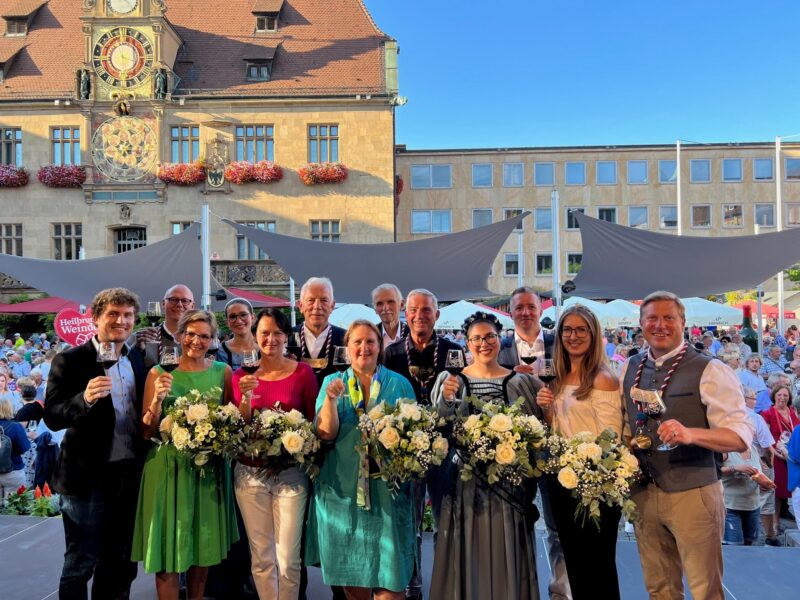 Gruppe von Menschen mit Blumensträußen und Gläsern vor einem historischen Gebäude.