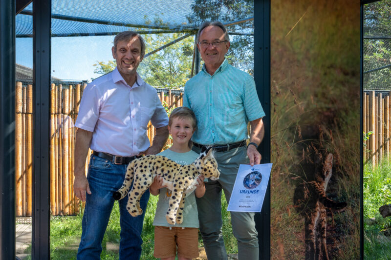 Drei Personen halten ein Stofftier und eine Urkunde in einem Tiergehege.