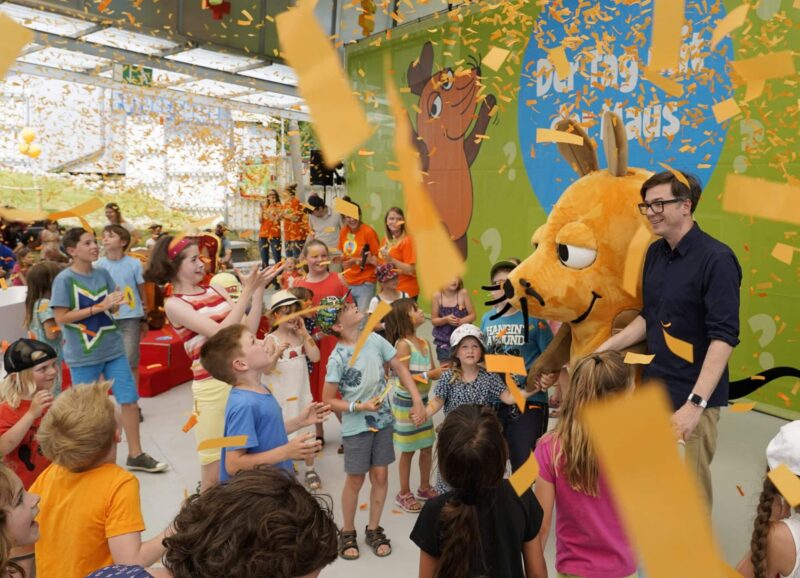 Kinder feiern mit einem Maskottchen und Konfetti in einem bunten Raum.