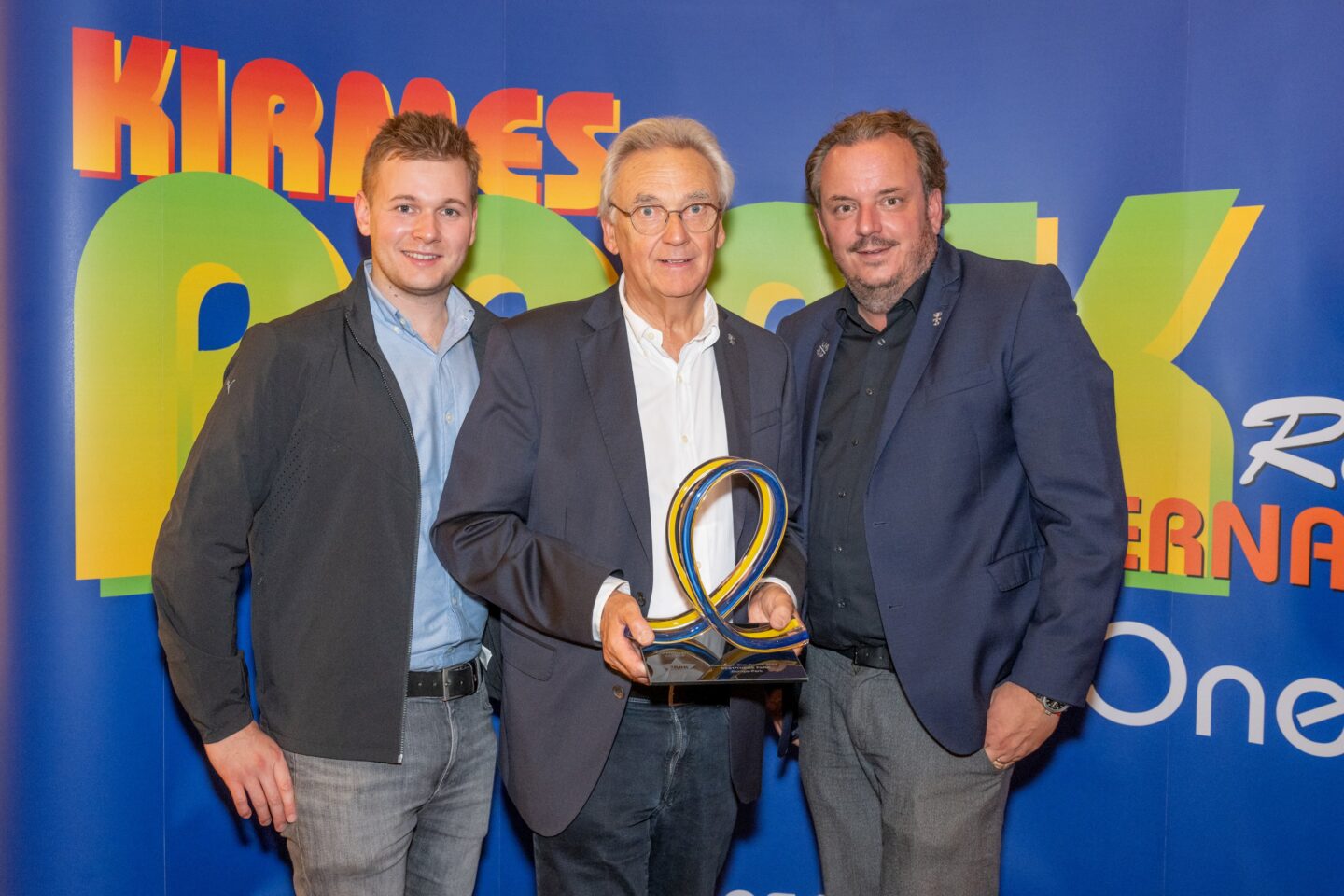 Drei Männer posieren mit einer Auszeichnung vor einem bunten Hintergrund.