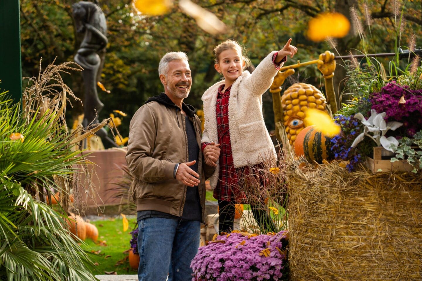 Ein Mann und ein Mädchen stehen in einem herbstlichen Garten mit bunten Blumen und Kürbissen.