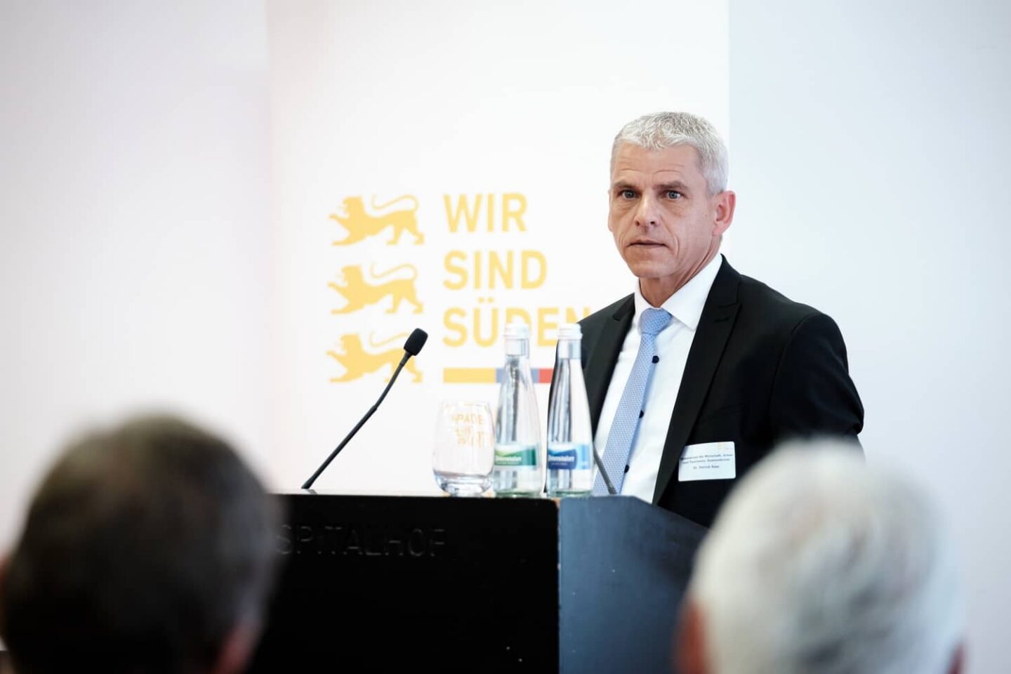 Mann im Anzug spricht an einem Rednerpult mit Wasserflaschen im Hintergrund.