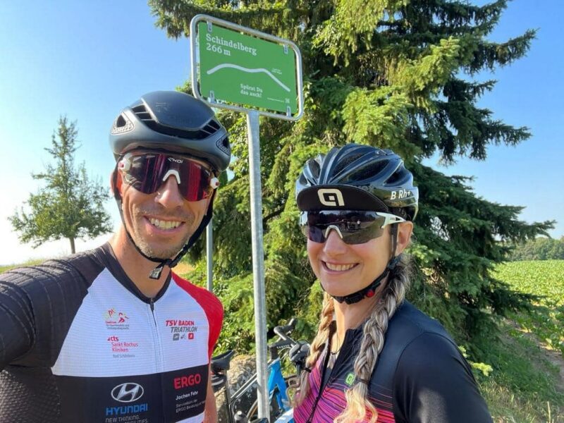 Zwei Radfahrer posieren vor einem Schild mit der Aufschrift 'Schindelberg 206 m'.