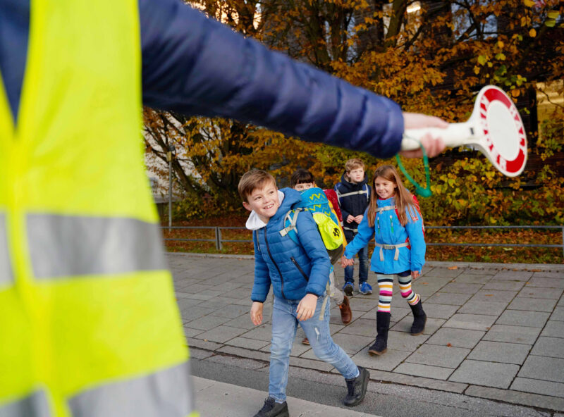 Kinder überqueren die Straße, während ein Verkehrsposten mit einer Haltescheibe signalisiert.