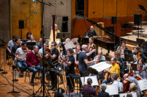 Orchesterprobe mit Musikern in einem Saal, verschiedene Instrumente und Notenständer sichtbar.