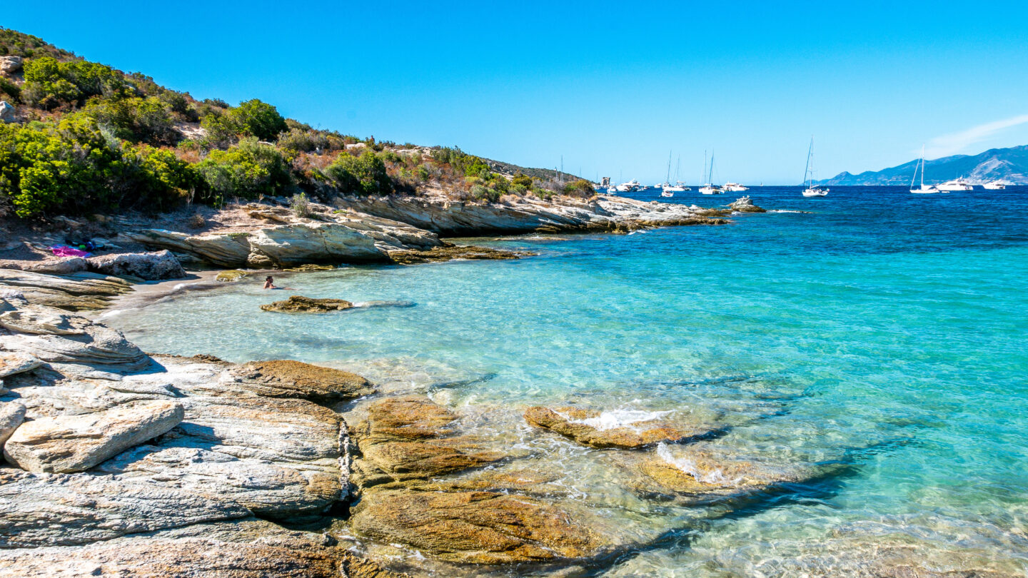 Klarer Strand mit Felsen und Yachten im Hintergrund.