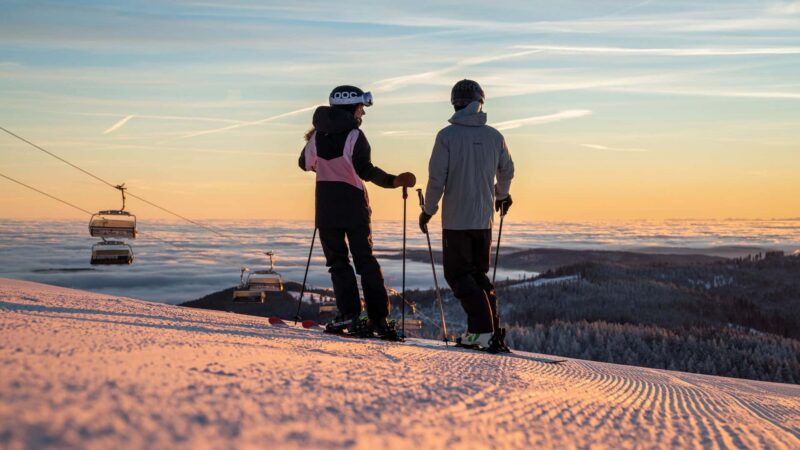Zwei Skifahrer stehen auf einer Piste bei Sonnenuntergang.
