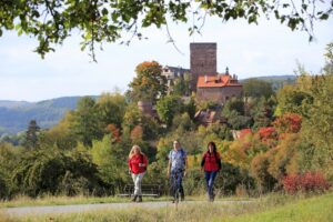 Drei Wanderer auf einem Weg mit einer Burg im Hintergrund und buntem Herbstlaub.
