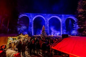 Weihnachtsmarkt unter einem beleuchteten Viadukt mit Menschen und einem Weihnachtsbaum.