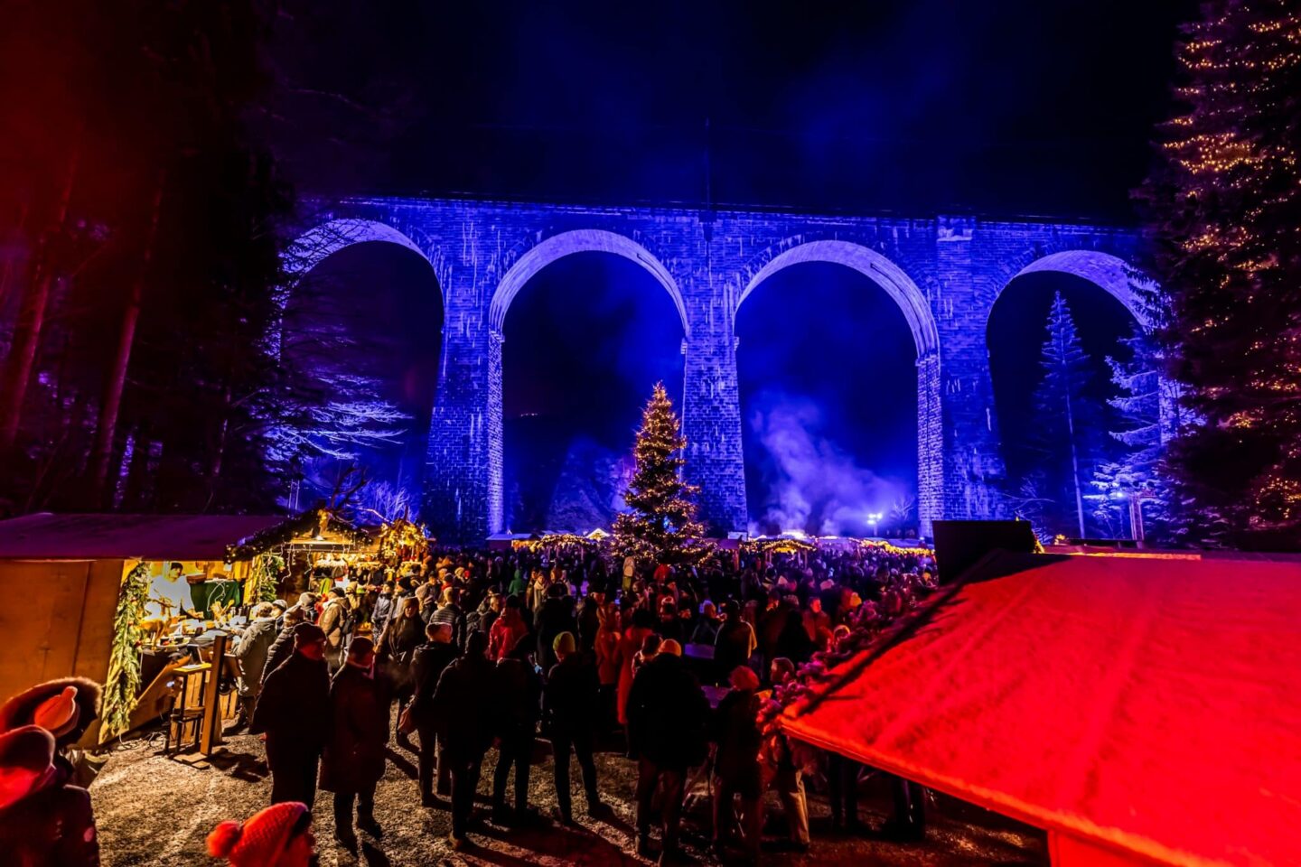 Weihnachtsmarkt unter einem beleuchteten Viadukt mit Menschen und einem Weihnachtsbaum.