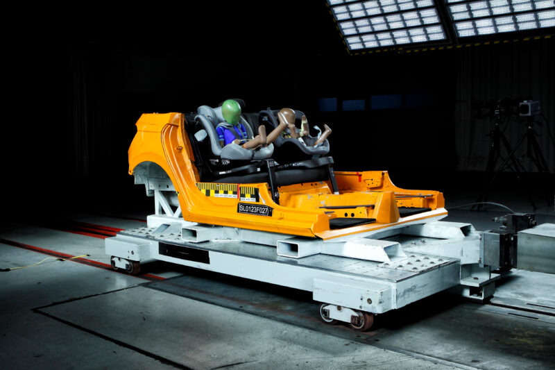 Testeinrichtung mit Dummy-Personen in einem gelben Fahrzeugrahmen.