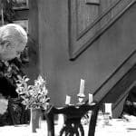 Schwarz-Weiß-Aufnahme aus „Dinner for One“: Butler James, ein älterer Herr mit grauem Haar und im klassischen Frack gekleidet, steht gebückt an der Seite von Miss Sophie, die an einer festlich gedeckten Tafel sitzt. Miss Sophie trägt ein elegantes Kleid mit Spitzenbesatz und lächelt höflich. Auf dem Tisch befinden sich mehrere Kerzenleuchter, Blumenarrangements und Geschirr. Im Hintergrund ist eine Holztreppe zu sehen, die zur Kulisse des berühmten Silvestersketches gehört.