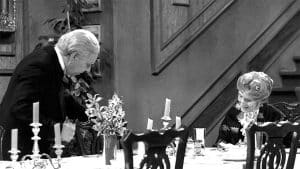 Schwarz-Weiß-Aufnahme aus „Dinner for One“: Butler James, ein älterer Herr mit grauem Haar und im klassischen Frack gekleidet, steht gebückt an der Seite von Miss Sophie, die an einer festlich gedeckten Tafel sitzt. Miss Sophie trägt ein elegantes Kleid mit Spitzenbesatz und lächelt höflich. Auf dem Tisch befinden sich mehrere Kerzenleuchter, Blumenarrangements und Geschirr. Im Hintergrund ist eine Holztreppe zu sehen, die zur Kulisse des berühmten Silvestersketches gehört.