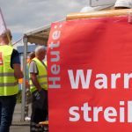 Ein rotes Schild mit der Aufschrift „Heute: Warnstreik!“ steht im Vordergrund. Im Hintergrund sind mehrere Personen in gelben Warnwesten mit ver.di-Logo zu sehen, die an einer Protestaktion teilnehmen. Einige Teilnehmer halten ver.di-Fahnen, während andere miteinander sprechen. Die Szene spielt sich auf einer Freifläche unter einem bewölkten Himmel ab.