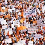 Große Menschenmenge von Ärztinnen und Ärzten bei einer Kundgebung, viele tragen weiße Kittel und orangefarbene Kappen. Sie halten Schilder und Banner mit Forderungen nach besseren Arbeitsbedingungen, fairen Gehältern und Work-Life-Balance hoch. Einige Schilder tragen Aufschriften wie 'Arbeitszeiterfassung JETZT!' und 'Wir streiken auch für unsere Patienten'. Die Veranstaltung wirkt gut organisiert, und die Teilnehmenden zeigen Entschlossenheit.
