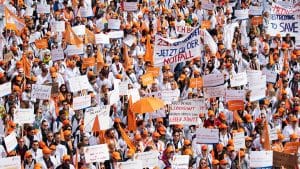 Große Menschenmenge von Ärztinnen und Ärzten bei einer Kundgebung, viele tragen weiße Kittel und orangefarbene Kappen. Sie halten Schilder und Banner mit Forderungen nach besseren Arbeitsbedingungen, fairen Gehältern und Work-Life-Balance hoch. Einige Schilder tragen Aufschriften wie 'Arbeitszeiterfassung JETZT!' und 'Wir streiken auch für unsere Patienten'. Die Veranstaltung wirkt gut organisiert, und die Teilnehmenden zeigen Entschlossenheit.