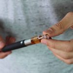 Nahaufnahme von Händen, die eine E-Zigarette halten und das Mundstück abnehmen. Die E-Zigarette enthält eine bernsteinfarbene Flüssigkeit im Tank. Der Hintergrund ist unscharf, die Person trägt ein graues T-Shirt.