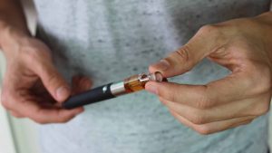 Nahaufnahme von Händen, die eine E-Zigarette halten und das Mundstück abnehmen. Die E-Zigarette enthält eine bernsteinfarbene Flüssigkeit im Tank. Der Hintergrund ist unscharf, die Person trägt ein graues T-Shirt.