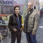 Das Ermittlerduo Cris Blohm (gespielt von Johanna Wokalek) und Dennis Eden (gespielt von Stephan Zinner) steht vor einem Tatort in einer bunten Künstlerkolonie, umgeben von Graffiti und Absperrband.