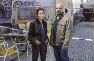 Das Ermittlerduo Cris Blohm (gespielt von Johanna Wokalek) und Dennis Eden (gespielt von Stephan Zinner) steht vor einem Tatort in einer bunten Künstlerkolonie, umgeben von Graffiti und Absperrband.