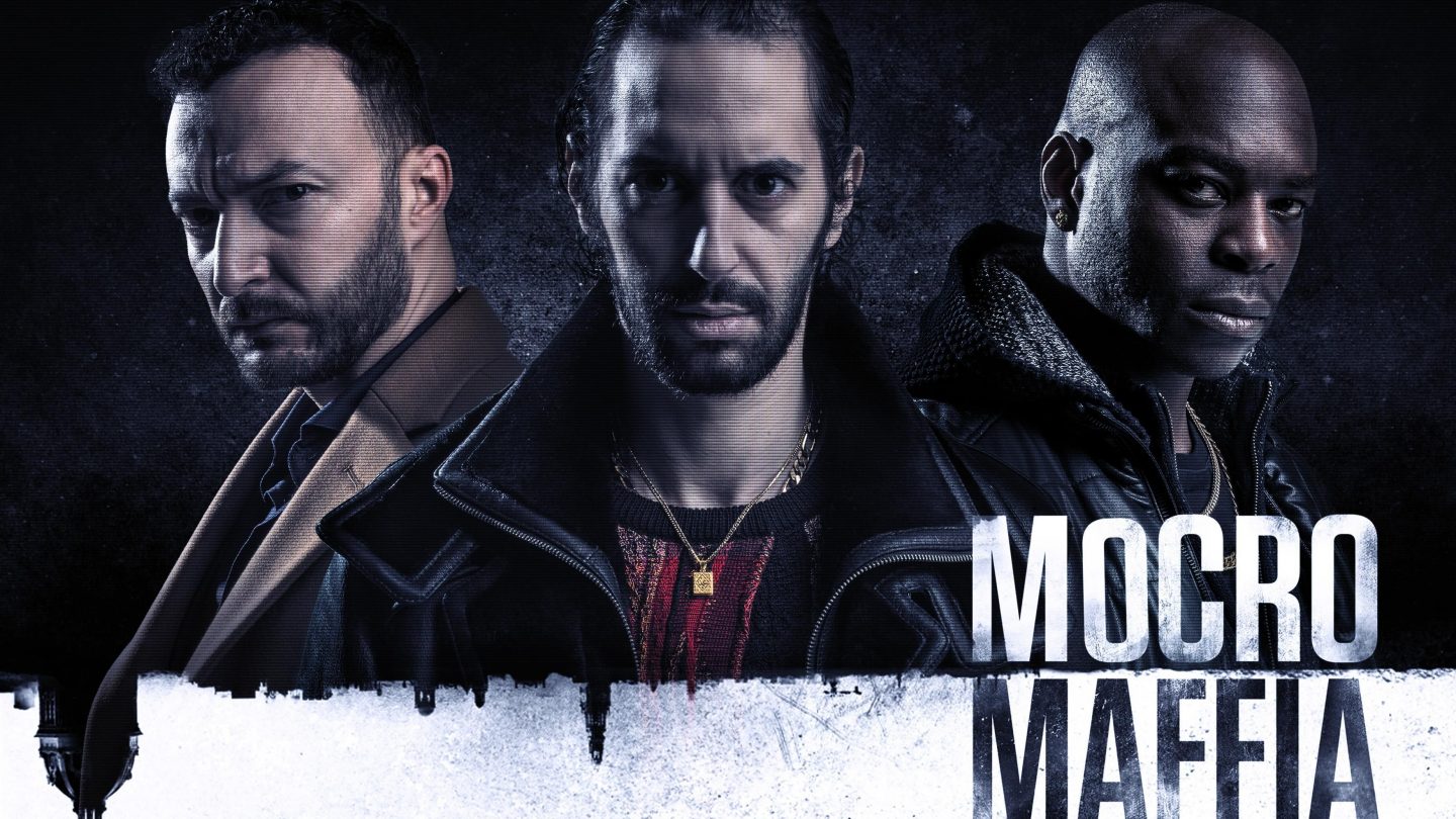 Promobild der Serie 'Mocro Maffia' mit drei Hauptcharakteren in dunkler Kleidung vor düsterem Hintergrund. Links ein Mann im braunen Mantel, in der Mitte ein Mann mit Lederjacke und ernstem Blick, rechts ein Mann mit rasiertem Kopf und schwarzer Jacke. Unter dem Bild steht der Titel 'Mocro Maffia' in großen weißen Buchstaben.
