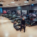 Lewis Hamilton sitzt lächelnd vor einer Reihe seiner sieben Weltmeister-Rennwagen im Mercedes-Benz Museum. Die ikonischen Formel-1-Boliden stehen in einer Reihe, während im Hintergrund Bilder von Hamiltons Karriere zu sehen sind.