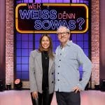 Negah Amiri, eine Stand-up-Comedienne und Moderatorin, steht neben Olli Dittrich, einem Schauspieler, Komiker und Musiker, im Studio von „Wer weiß denn sowas?“. Im Hintergrund leuchtet das Logo der Quizshow in auffälligen Neonfarben, eingerahmt von einer Backsteinwand und Studiobeleuchtung. Beide lächeln in die Kamera und vermitteln eine lockere, freundliche Atmosphäre, passend zum humorvollen Charakter der Show.