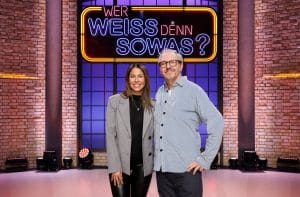 Negah Amiri, eine Stand-up-Comedienne und Moderatorin, steht neben Olli Dittrich, einem Schauspieler, Komiker und Musiker, im Studio von „Wer weiß denn sowas?“. Im Hintergrund leuchtet das Logo der Quizshow in auffälligen Neonfarben, eingerahmt von einer Backsteinwand und Studiobeleuchtung. Beide lächeln in die Kamera und vermitteln eine lockere, freundliche Atmosphäre, passend zum humorvollen Charakter der Show.