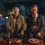 Szene aus dem „Tatort: Man stirbt nur zweimal“ mit Axel Prahl als Hauptkommissar Frank Thiel (links) und Jan Josef Liefers als Professor Karl-Friedrich Boerne (rechts). Beide sitzen abends an einem Tisch im Freien, vor sich Teller mit Pizza und ein Glas Rotwein. Thiel trägt lässige Kleidung, während Boerne im Anzug und Krawatte formell gekleidet ist. Die Stimmung ist angespannt und mysteriös, untermalt von warmer, künstlicher Beleuchtung. Im Hintergrund ist ein Garten mit Lampen zu sehen.
