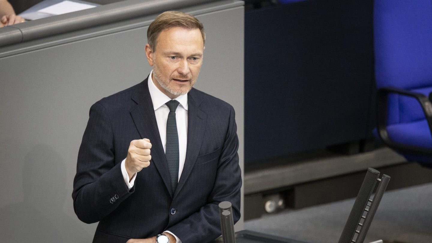 Christian Lindner, der FDP-Vorsitzende, steht am Rednerpult im Bundestag. Er trägt einen dunkelblauen Anzug mit weißem Hemd und dunkler Krawatte. Mit entschlossenem Gesichtsausdruck und geballter Faust hält er eine Rede. Vor ihm liegt ein Redemanuskript und ein Glas Wasser. Im Hintergrund sind die Sitzplätze des Bundestags sichtbar.