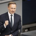 Christian Lindner, der FDP-Vorsitzende, steht am Rednerpult im Bundestag. Er trägt einen dunkelblauen Anzug mit weißem Hemd und dunkler Krawatte. Mit entschlossenem Gesichtsausdruck und geballter Faust hält er eine Rede. Vor ihm liegt ein Redemanuskript und ein Glas Wasser. Im Hintergrund sind die Sitzplätze des Bundestags sichtbar.