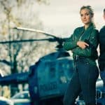 Ein dynamisches Duo aus der Serie „Alarm für Cobra 11 – Die Autobahnpolizei“ posiert vor einem Polizeihubschrauber. Die beiden Charaktere, gespielt von Pia Stutzenstein (links) und Erdoğan Atalay (rechts), tragen lässige Polizeikleidung und halten Waffen in den Händen, bereit für die nächste actionreiche Mission. Im Hintergrund stehen Polizeiautos und der Hubschrauber, die die Szenerie