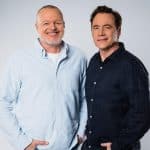 „Stefan Raab und Michael 'Bully' Herbig stehen nebeneinander vor einem neutralen Hintergrund. Raab trägt ein hellblaues Hemd und lächelt freundlich, während Herbig ein dunkles Hemd trägt und dezent lächelt.“