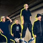 Die Fantastischen Vier posieren in einem urbanen, industriellen Setting. Die Mitglieder sind mit auffälligen gelben Konturen hervorgehoben, während sie lässige Kleidung tragen. Die Szene vermittelt eine moderne, dynamische Atmosphäre und unterstreicht ihren ikonischen Status in der Musikszene.
